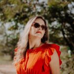 Elegant woman in sunglasses and red dress poses outdoors on a sunny summer day.